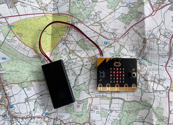 una micro:bit i una bateria adjunta que es recolzen a la part superior d'un mapa amb una fletxa cap amunt que es mostra a la pantalla LED de la micro:bit 